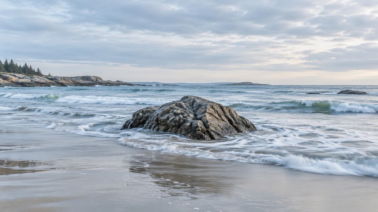 A single rock standing firm in the shallows as waves break around it on an overcast northern beach